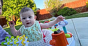 Jenna is registered to the contest to win money with this photo: activity_walker, baby, bib, child, expression, face, fence, grass, greenery, happy, infant, nature, outdoor, person, play, sidewalk, summer, sunlight, toys, young