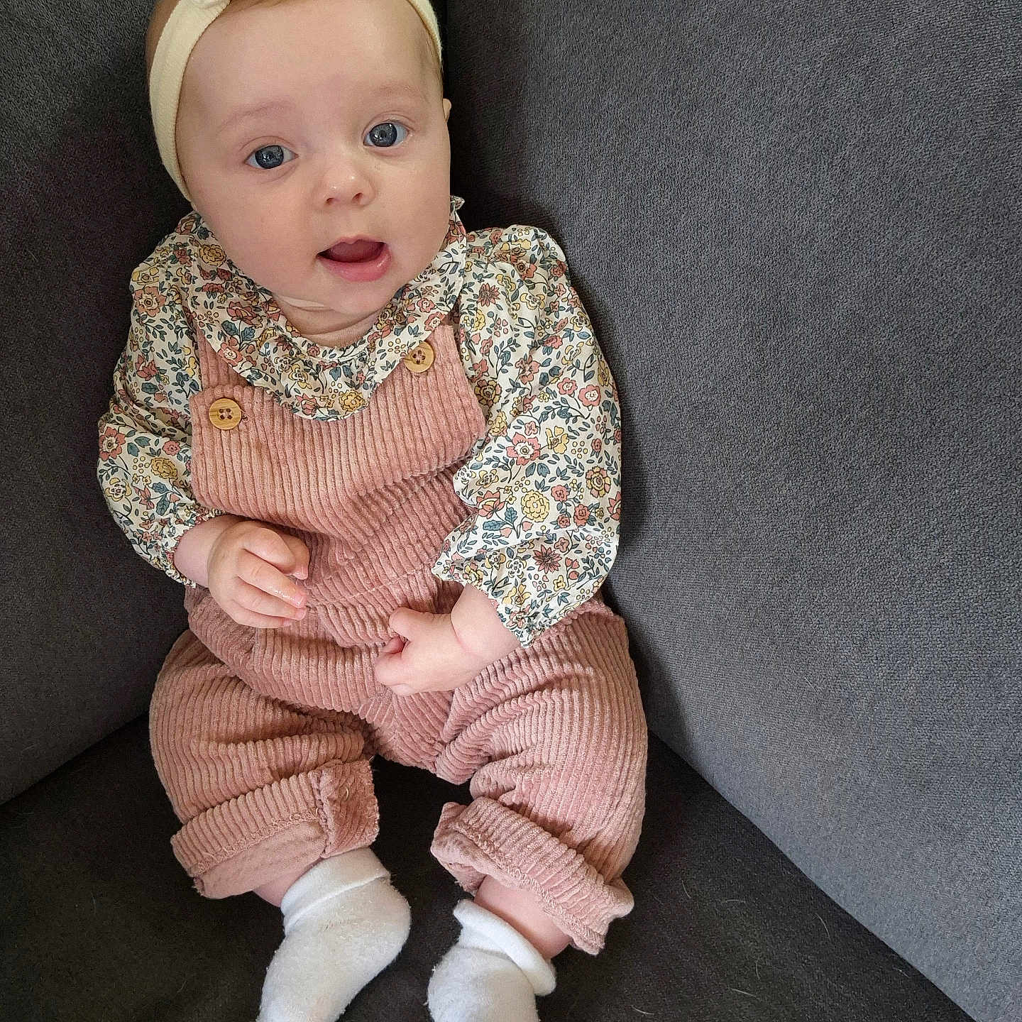 Capucine participe au concours pour gagner de l'argent avec cette photo : baby, blue_eyes, child, comfortable, couch, cozy, cute, fashion, floral_shirt, headband, indoors, infant, person, pink_overalls, portrait, seated, smiling, socks, soft_lighting, young_child
