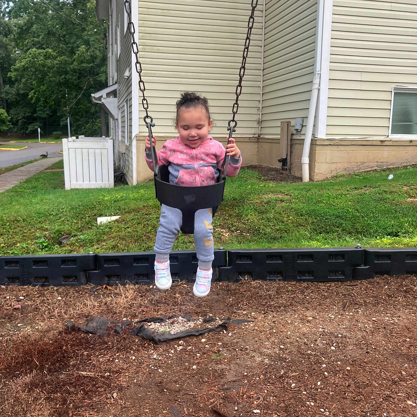 Xaviona joined the competition — help win amazing prizes! child, toddler, swing, playground, outdoor, smiling, happy, sitting, swinging, pink_jacket, gray_pants, sneakers, chains, backyard, house, grass, trees, mulch, portrait, playtime