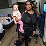 Xaviona is registered to the contest to win money with this photo: child, toddler, adult, caregiver, woman, holding, indoor, bedroom, standing, portrait, family, pink_pajamas, black_clothing, glasses, smiling, home, television, stroller, bed, wood_floor