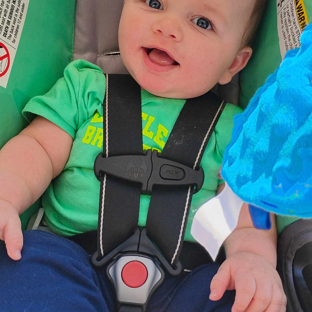 Karson is registered to the contest to win money with this photo: baby, child, car_seat, green_shirt, blue_pants, smiling, happy, infant, seatbelt, toy, blue_toy, cute, portrait, sitting, foot, hand, indoor, safety, young_child, face