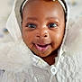 baby, smiling, close_up, white_clothing, headband, flower, hood, knitwear, happy, infant, cute, portrait, child, face, eyes, buttoned_clothing, soft_texture, indoor, warm, adorable