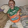 child, toddler, smiling, sitting, indoor, bed, blanket, plush_toy, elephant, green_shirt, orange_bowl, blue_liquid, messy, playful, portrait, home, furniture, face, short_hair, happy