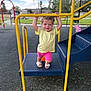 child, toddler, playground, yellow_bars, smiling, outdoor, play_equipment, sandals, pink_shorts, yellow_shirt, steps, blue_play_structure, fence, grass, cloudy_sky, happy, person, summer, fun, recreation