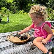 Blaire is registered to the contest to win money with this photo: toddler, child, curly_hair, pink_outfit, sandals, stones, wooden_table, outdoor, grass, garden, statue, sunny, nature, playing, summer, daylight, greenery, concentration, park, casual
