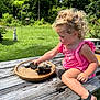 toddler, child, curly_hair, pink_outfit, sandals, stones, wooden_table, outdoor, grass, garden, statue, sunny, nature, playing, summer, daylight, greenery, concentration, park, casual