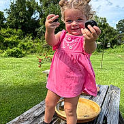 Blaire is registered to the contest to win money with this photo: toddler, child, girl, pink_dress, curly_hair, smile, outdoor, grass, trees, wooden_table, stones, sandals, sunlight, nature, playful, portrait, summer, greenery, happy, standing