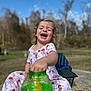 child, toddler, girl, laughing, curly_hair, princess_pajamas, bubbles, bubble_solution, green_jar, outdoor, blue_sky, blurred_background, chair, sitting, play, hand, smile, portrait, candid, toy