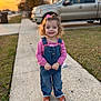 child, toddler, girl, smile, curly_hair, overalls, pink_shirt, sandals, sidewalk, suburban, pickup_truck, sunset, grass, pavement, portrait, outdoor, happy, front_yard, bokeh, candid
