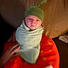 newborn, baby, infant, swaddled, green_hat, blanket, red_blanket, polka_dots, portrait, face, serious_expression, indoors, warm_lighting, closeup, hand, adult_hand, cuddling, baby_clothing, cozy, family