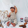 child, toddler, baby, chef_hat, baking, cookie, jam_jar, mason_jar, chocolate_milk, eggs, rolling_pin, cutting_board, number_ten, white_sheet, sitting, bare_feet, romper, snack, kitchen_prop, portrait