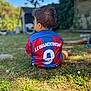 Nahel a rejoint le concours — aidez-le/la à gagner de superbes lots ! child, toddler, back_view, football_jersey, grass, outdoor, garden, daylight, nature, greenery, casual_clothing, short_hair, sportswear, number_9, blue, red, playing, sitting, sunny, blurred_background