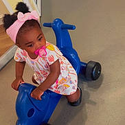 Nylah joined the competition — help win amazing prizes! toddler, child, pacifier, headband, bow, ride_on_toy, blue_toy, indoor, dress, floral_pattern, black_shoes, floor, curious, playing, person, young_child, plastic_toy, cute, baby, pigtails