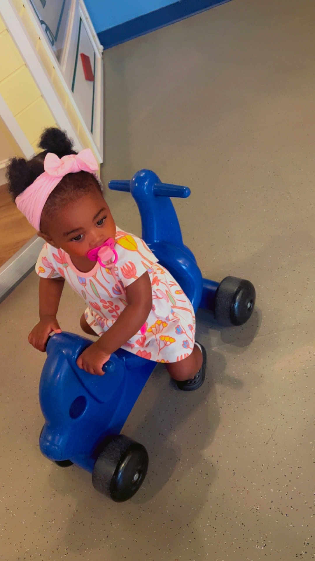 Nylah joined the competition — help win amazing prizes! toddler, child, pacifier, headband, bow, ride_on_toy, blue_toy, indoor, dress, floral_pattern, black_shoes, floor, curious, playing, person, young_child, plastic_toy, cute, baby, pigtails