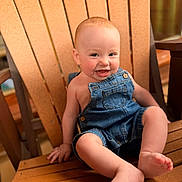 Ronnie is registered to the contest to win money with this photo: baby, child, denim_overalls, smiling, happy, wooden_chair, furniture, indoor, person, cute, portrait, skin, hand, foot, blue_nails, baby_clothing, sitting, infant, young_child, expression