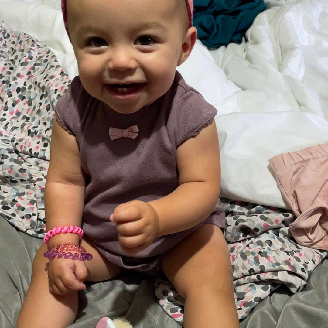 Jasmine Yacoub participe au concours pour gagner de l'argent avec cette photo : accessories, baby, baby_brush, bed, blanket, bracelets, child, cute, face, happy, headband, indoors, infant, person, pink_socks, purple_clothing, sitting, skin, smiling, toy