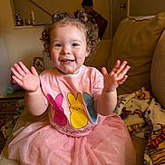 Blakely joined the competition — help win amazing prizes! toddler, child, girl, pink_dress, curly_hair, clapping, couch, blanket, toy, indoor, smiling, happy, person, home, play, background, furniture, casual_clothing, family, curly