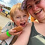 child, woman, smiling, donut, food, outdoor, selfie, happy, face, hat, green_wristband, casual_clothing, daylight, blue_sky, clouds, playful, fun, park, candid, people