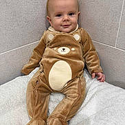 Jürgen a rejoint le concours — aidez-le/la à gagner de superbes lots ! baby, child, onesie, bear_costume, smile, face, infant, cuteness, tiled_wall, blanket, soft_texture, seated, indoors, cozy, young_child, happy, small_hands, footed_pajamas, wall_corner, cute