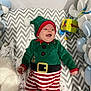 baby, infant, smiling, christmas_outfit, elf_costume, striped_pants, crib, blanket, toy, soft_cushions, happy, holiday, festive, red_and_green, plush, cute, child, indoors, cheerful, lying_down