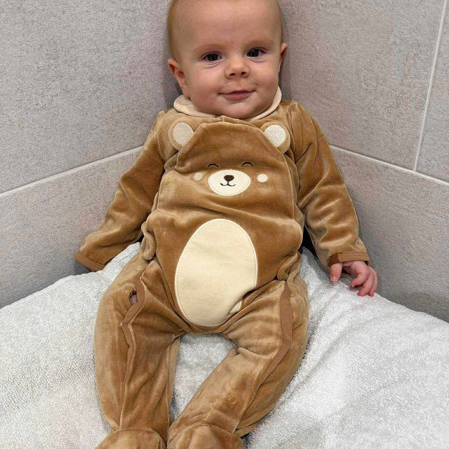 Jürgen a rejoint le concours — aidez-le/la à gagner de superbes lots ! baby, bear_costume, blanket, child, cozy, cute, cuteness, face, footed_pajamas, happy, indoors, infant, onesie, seated, small_hands, smile, soft_texture, tiled_wall, wall_corner, young_child