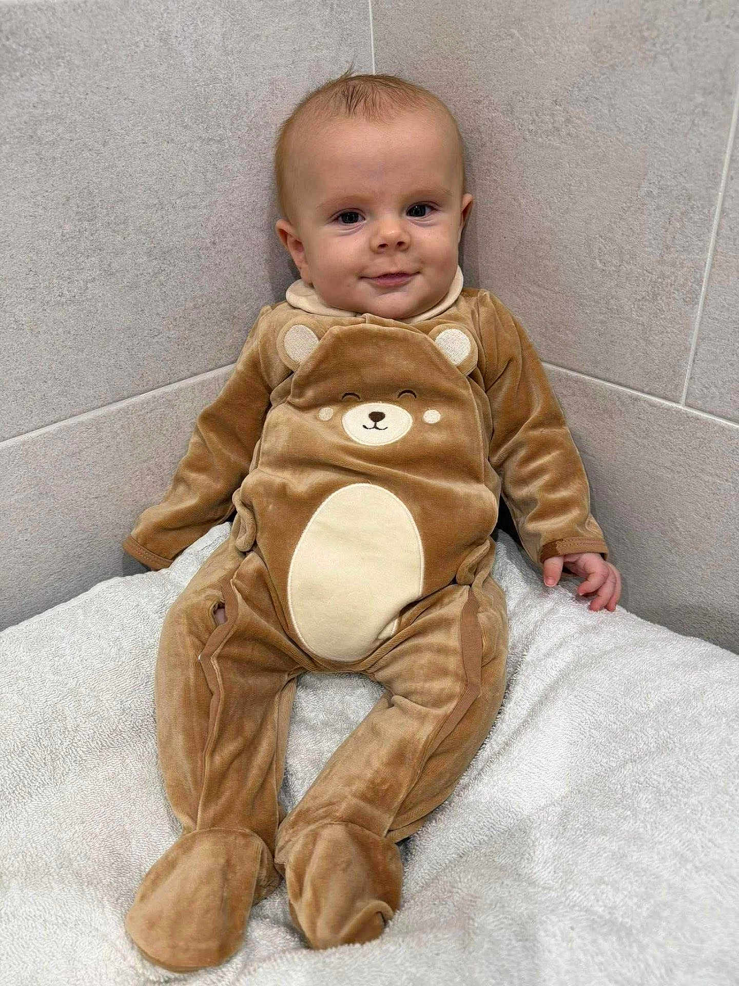 Jürgen a rejoint le concours — aidez-le/la à gagner de superbes lots ! baby, child, onesie, bear_costume, smile, face, infant, cuteness, tiled_wall, blanket, soft_texture, seated, indoors, cozy, young_child, happy, small_hands, footed_pajamas, wall_corner, cute