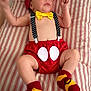 baby, infant, child, red_hat, yellow_bow_tie, polka_dot_suspender, red_shorts, striped_bedspread, socks, mickey_mouse_theme, lying_down, raised_arms, cute, indoor, portrait, clothing, baby_fashion, soft_lighting, human, young_child