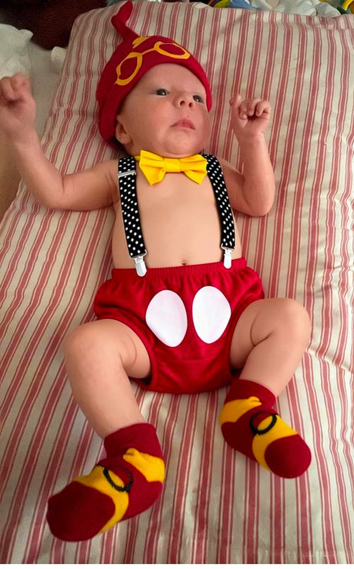 Thomas joined the competition — help win amazing prizes! baby, infant, child, red_hat, yellow_bow_tie, polka_dot_suspender, red_shorts, striped_bedspread, socks, mickey_mouse_theme, lying_down, raised_arms, cute, indoor, portrait, clothing, baby_fashion, soft_lighting, human, young_child