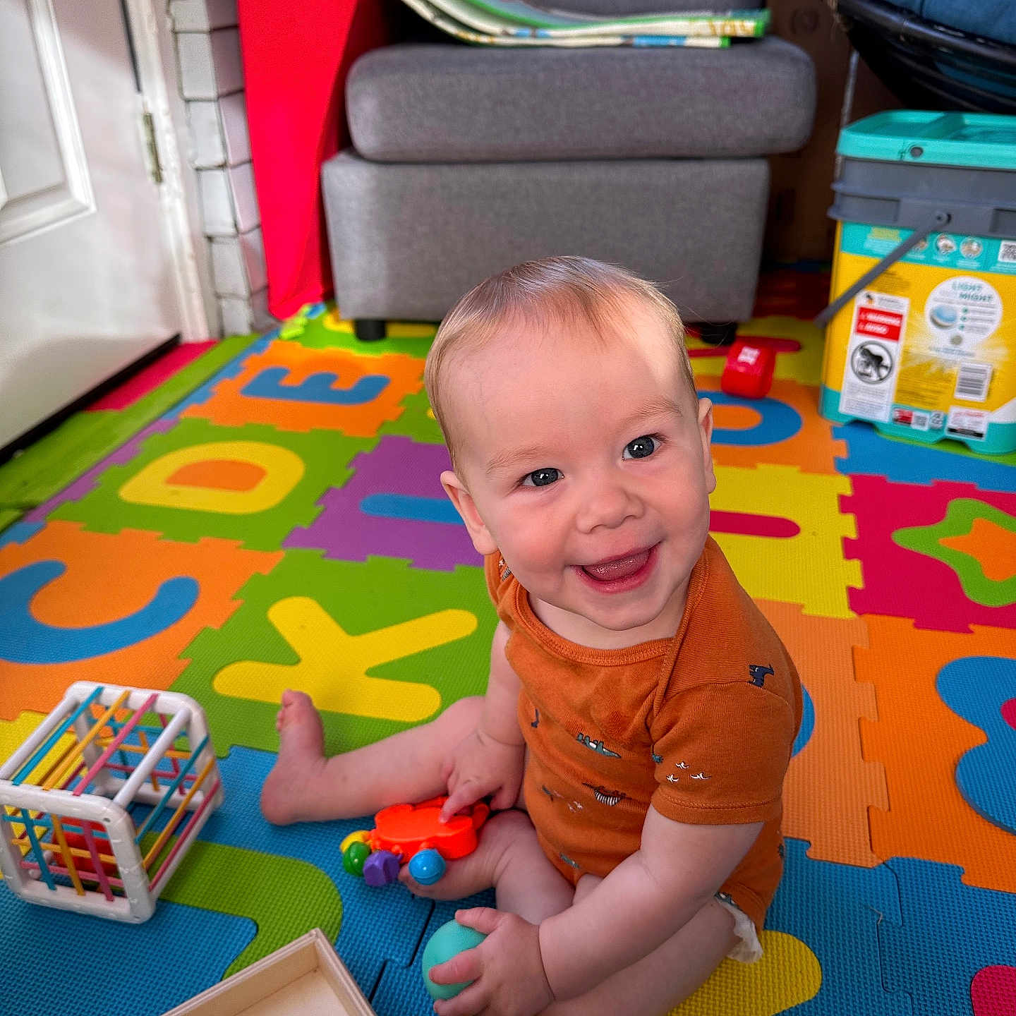 Liam joined the competition — help win amazing prizes! baby, bright, child, colorful, cube_toy, cute, floor, foam_mat, happy, indoor, infant, living_room, orange_clothing, person, play, playtime, sitting, smiling, toy, wooden_box