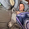 adult, bed, blanket, cat, comfort, cozy, facial_hair, furniture, gray_cat, home, indoor, man, nap, pillow, relaxation, rest, sleep, sleeping, stretching, tattoo