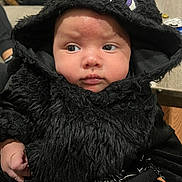 Emerik is registered to the contest to win money with this photo: baby, child, costume, black_fur, hood, cute, face, expression, indoor, person, hand, purple_eyes, clothing, portrait, curious, soft_texture, tattoo, background, table, can