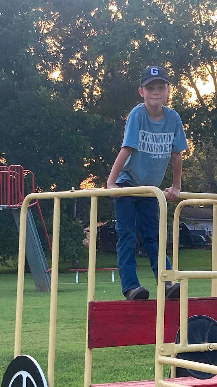 Skyler is registered to the contest to win money with this photo: baseball_cap, cap, city, electric_blue, flash_photography, fun, grass, handrail, hat, headwear, jeans, joy, leisure, outdoor_play_equipment, person, plant, play, recreation, shorts, sitting