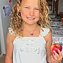 child, girl, smiling, curly_hair, blonde_hair, white_top, blue_shorts, apple, indoor, kitchen, clock, staircase, table, tablecloth, wooden_floor, necklace, happy, person, casual_clothing, holding_object