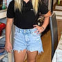child, girl, blonde_hair, smiling, standing, black_polo_shirt, denim_shorts, white_sneakers, indoor, kitchen, wood_floor, plant, casual_clothing, young_person, happy, portrait, home_interior, furniture, hand_on_hip, natural_light