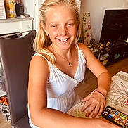 Chelsea a rejoint le concours — aidez-le/la à gagner de superbes lots ! child, girl, smiling, blonde_hair, white_dress, sitting, table, smartphone, chair, indoor, home, decorations, clock, wooden_floor, face, hand, necklace, earrings, happy, casual