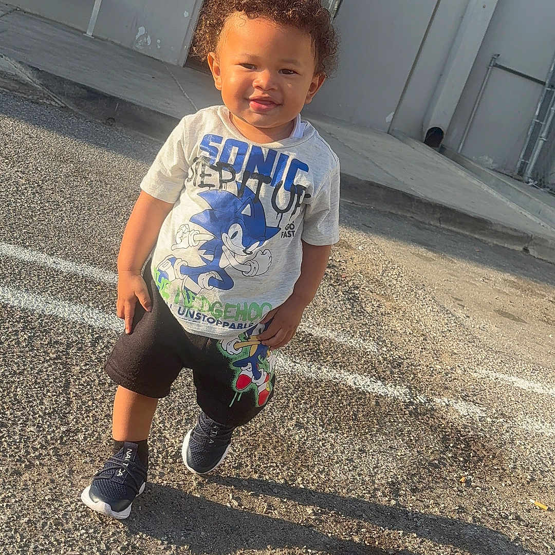 Ryan joined the competition — help win amazing prizes! child, toddler, boy, sonic_the_hedgehog, tshirt, shorts, sneakers, outdoor, parking_lot, shadow, sunlight, curly_hair, building, bench, concrete, sidewalk, smiling, standing, daytime, casual_clothing