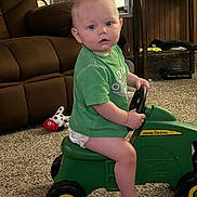 Liam is registered to the contest to win money with this photo: toddler, child, indoor, toy, ride_on_toy, john_deere, green_shirt, carpet, living_room, furniture, plush_toy, brown_couch, cabinet, steering_wheel, barefoot, curious, seated, carpeted_floor, diaper, face