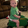 toddler, child, indoor, toy, ride_on_toy, john_deere, green_shirt, carpet, living_room, furniture, plush_toy, brown_couch, cabinet, steering_wheel, barefoot, curious, seated, carpeted_floor, diaper, face