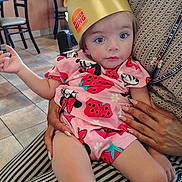 Emerson is registered to the contest to win money with this photo: toddler, child, burger_king_crown, pink_clothing, strawberry_pattern, sitting, indoor, restaurant, person_holding_child, blue_eyes, curious_expression, hand, striped_pants, tile_floor, chair, table, casual, cute, face, portrait