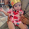 toddler, child, burger_king_crown, pink_clothing, strawberry_pattern, sitting, indoor, restaurant, person_holding_child, blue_eyes, curious_expression, hand, striped_pants, tile_floor, chair, table, casual, cute, face, portrait