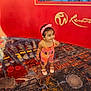 Veena joined the competition — help win amazing prizes! toddler, child, colorful_clothing, headband, pink, smiling, standing, indoor, carpet, patterned_carpet, red_wall, logo, playful, cute, person, young_child, earrings, footwear, happy, portrait