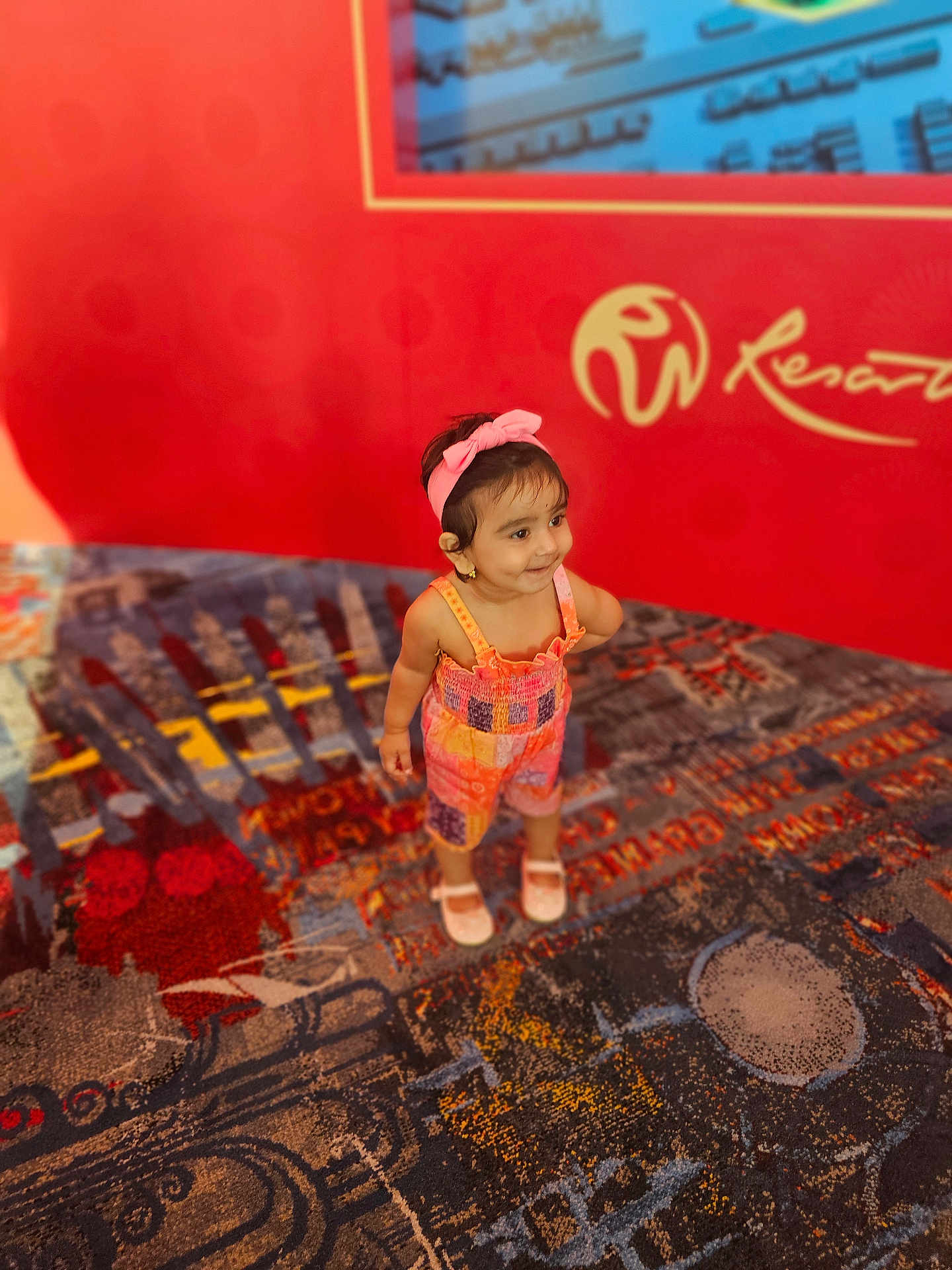 Veena joined the competition — help win amazing prizes! toddler, child, colorful_clothing, headband, pink, smiling, standing, indoor, carpet, patterned_carpet, red_wall, logo, playful, cute, person, young_child, earrings, footwear, happy, portrait