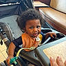 toddler, child, stroller, pacifier, afro_hair, indoor, table, hand, curious, orange_bib, black_shirt, seat, diner, tray, floor_tiles, person, face, expression, baby, furniture