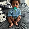 baby, pacifier, bed, blanket, pillow, blue_shirt, baby_clothing, indoor, child, curious, sitting, cross_legged, hair, skin, foot, hand, person, portrait, young_child, cozy