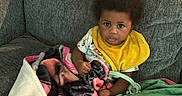 Akyrie joined the competition — help win amazing prizes! baby, blanket, child, colorful, couch, cozy, curly_hair, cute, furniture, home, indoor, infant, looking_at_camera, person, portrait, relaxed, seated, soft_texture, yellow_bib, young_child