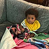 Akyrie joined the competition — help win amazing prizes! baby, child, infant, couch, blanket, yellow_bib, indoor, furniture, curly_hair, person, seated, colorful, cozy, portrait, cute, young_child, looking_at_camera, soft_texture, home, relaxed