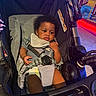 baby, child, stroller, seatbelt, diaper, face, curly_hair, indoor, lighting, hand, clothing, comfort, black_hair, expression, infant, photography, seat, baby_bib, person, cute