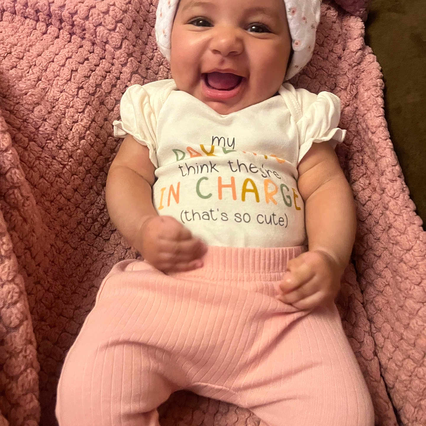 Nova is registered to the contest to win money with this photo: apparel, baby, blanket, child, cute, face, happy, hat, headwear, indoors, infant, lying_down, person, pink_pants, portrait, smiling, soft, texture, white_shirt, young_child