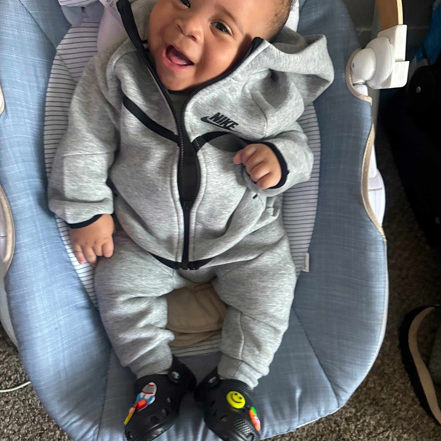 Jermoni is registered to the contest to win money with this photo: baby, smiling, infant, tracksuit, nike, baby_rocker, toy, stuffed_animal, black_shoes, colorful_patches, carpet, happy, child, indoor, casual_clothing, cute, portrait, person, seated, cozy