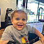toddler, child, smiling, happy, curly_hair, blue_eyes, high_chair, indoor, restaurant, table, casual_clothing, short_sleeve_shirt, daylight, window, floor_tiles, person, portrait, seated, furniture, young_child